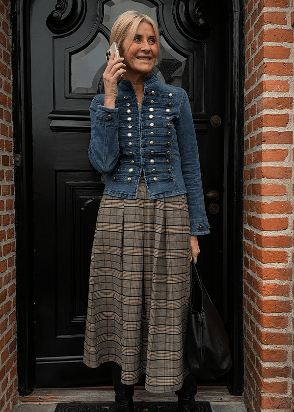 En kvinde står foran en sort dør og taler i mobiltelefonen. Hun er iført en militærinspireret jakke i denim, en midi-nederdel i ternet stof og en skuldertaske.