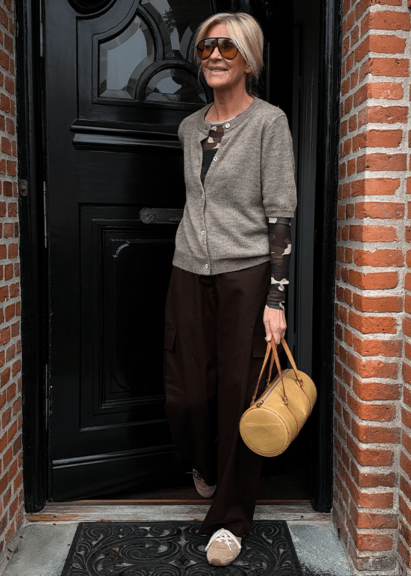 En kvinde iført en beige cardigan over en mønstret bluse, mørke bukser og store solbriller står i en døråbning og holder en beige, cylindrisk håndtaske. - Mandags outfittet
