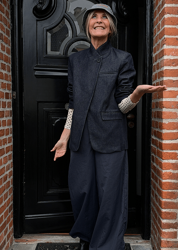En kvinde iført en bucket hat, en mørk blazer og bukser med brede ben står foran en sort dør og gestikulerer med hænderne.