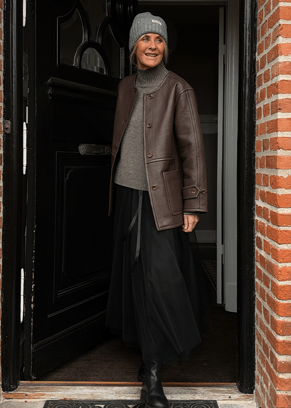 En kvinde iført en grå hue, en brun jakke, en grå trøje og en sort nederdel står i en åben døråbning. - Torsdags outfittet