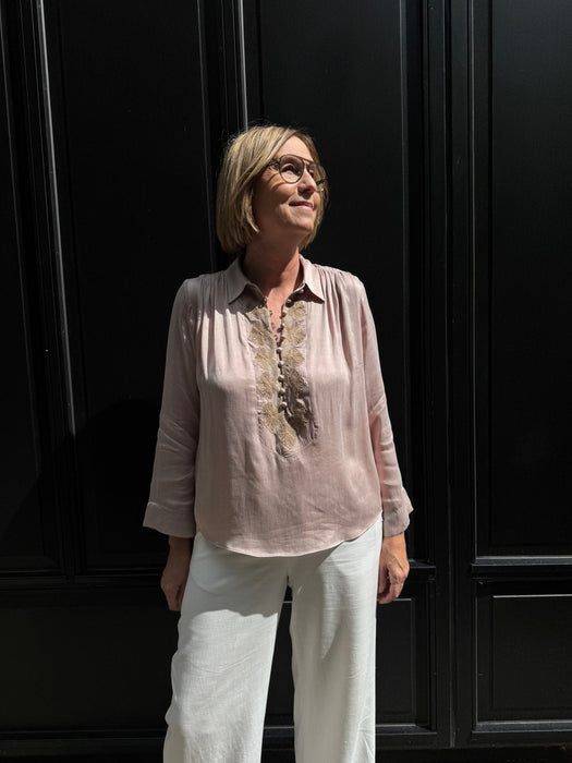 En kvinde med briller, hvide bukser og en bluse fra Gustav Sikka Lysebrun nyder solskinnet ved en elegant sort væg. - Rosa skjorte og hvide bukser