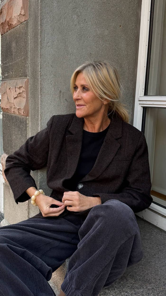 En kvinde med blondt hår sidder på gulvet ved et vindue iført en mørk blazer, en sort skjorte og grå fløjlsbukser. Hun kigger til siden med et let smil. - Blazer efterår