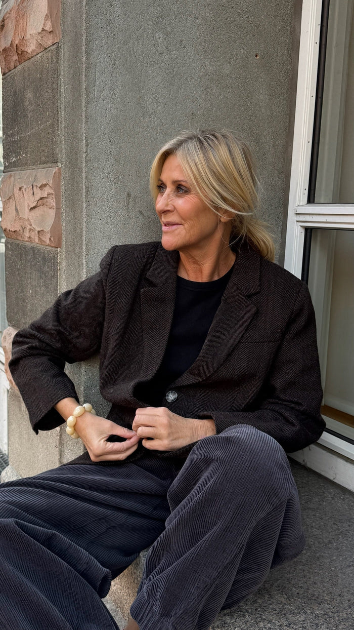 En kvinde med blondt hår sidder på gulvet ved et vindue iført en mørk blazer, en sort skjorte og grå fløjlsbukser. Hun kigger til siden med et let smil. - Blazer efterår