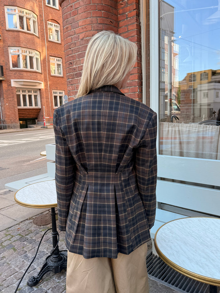 En person med langt blondt hår, iført en brun ternet blazer og beige bukser, står udenfor ved nogle runde caféborde på et brostensbelagt fortov og har ryggen til kameraet. - Ternet blazer