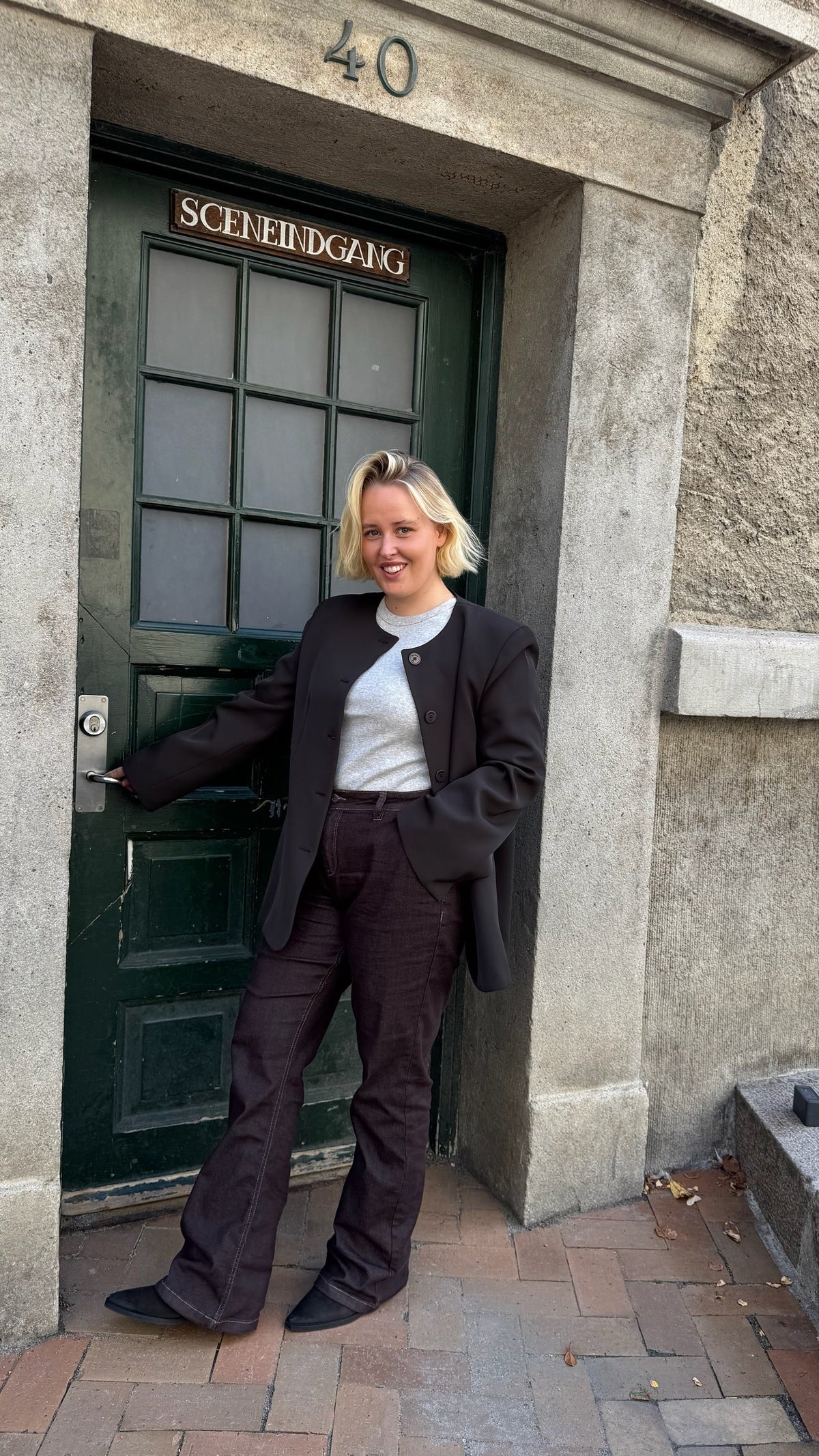 En person med blondt hår står foran en grøn dør med skiltet »SCENEINDGANG«. Personen har en mørk blazer, en grå skjorte og mørke bukser på og smiler til kameraet med den ene hånd på dørhåndtaget.