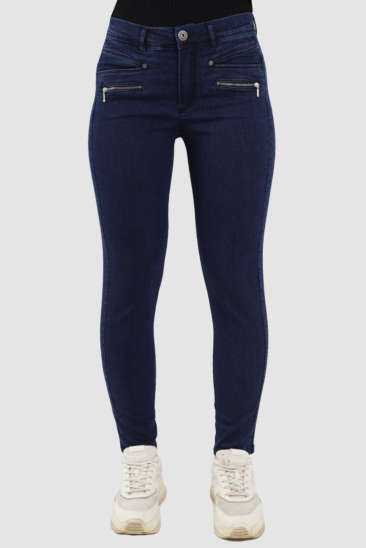 En person iført 2-Biz Rany Mørk-skinnyjeans i denim og hvide sneakers, der står foran en ensfarvet baggrund.