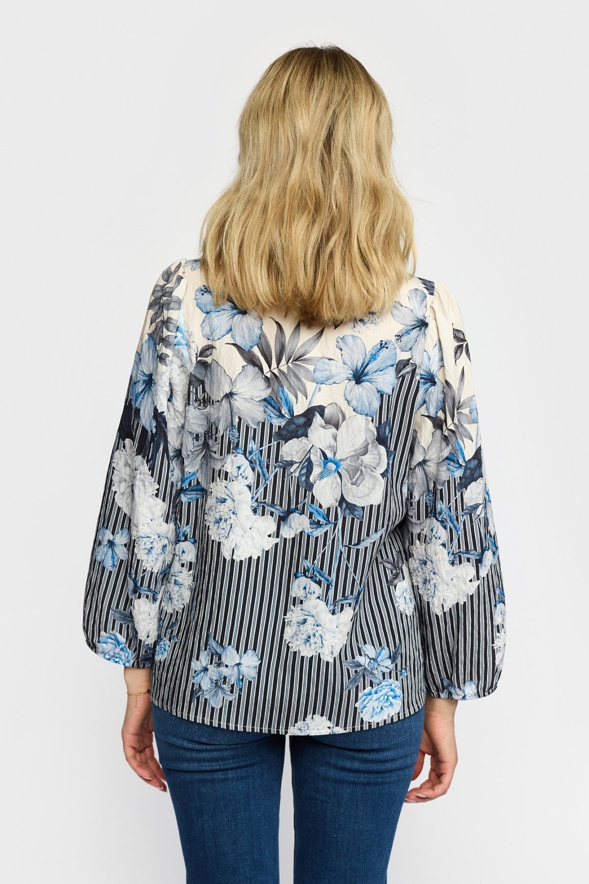 Kvinde, der står med ryggen til, iført en mønstret bluse fra 2-Biz Jana i blå og mørke jeans, foran en ensfarvet baggrund.