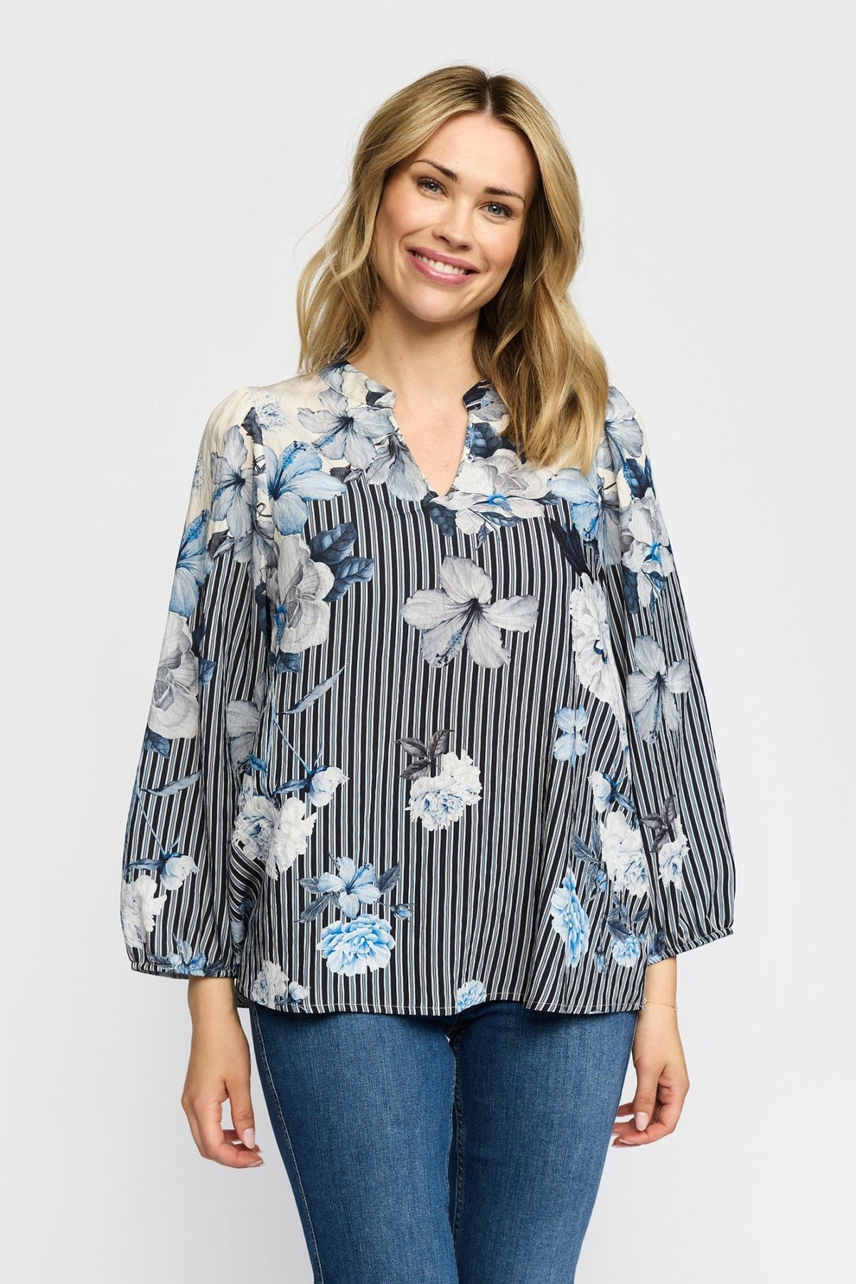 En kvinde har en mønstret bluse fra 2-Biz Jana Blå og blå jeans på; hun står og smiler til kameraet.