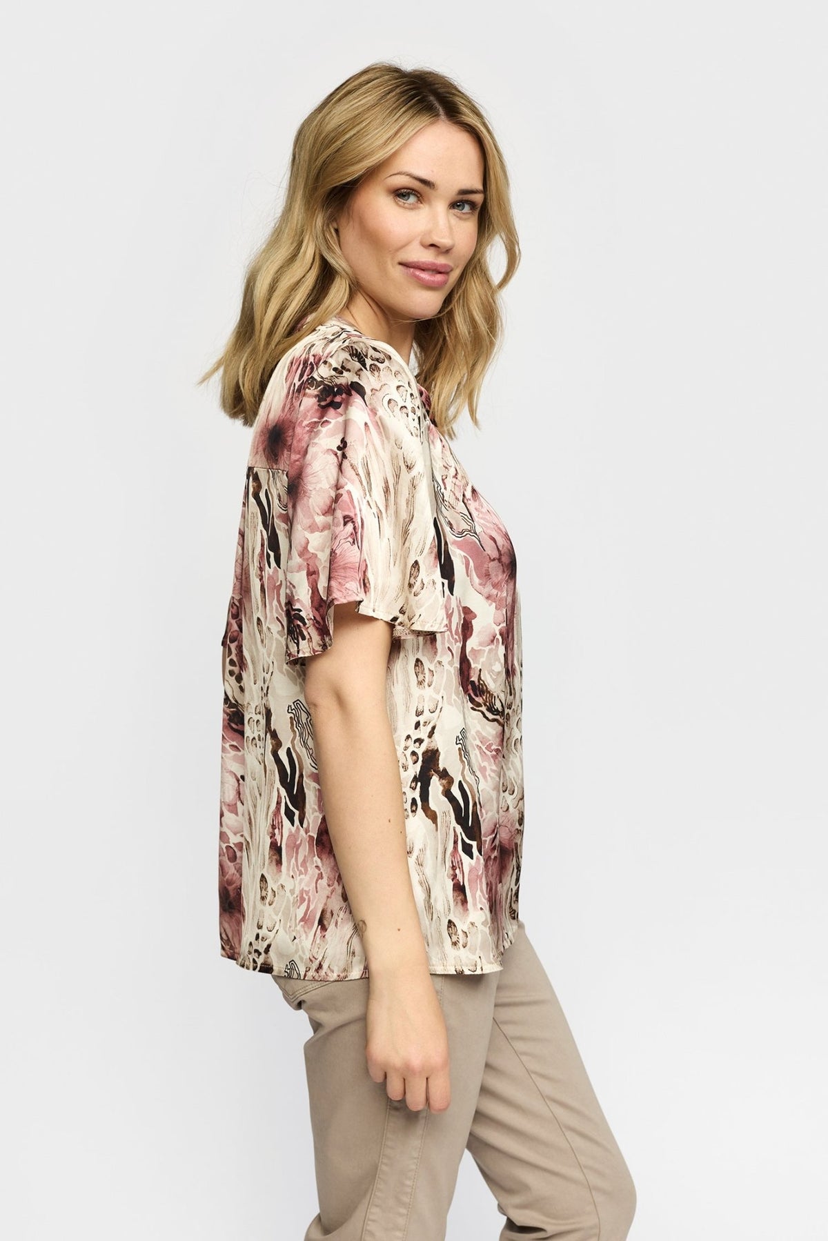 Kvinde iført en 2-Biz Amora Rosa-mønstret bluse og beige bukser, der står med siden til et ensfarvet baggrund.