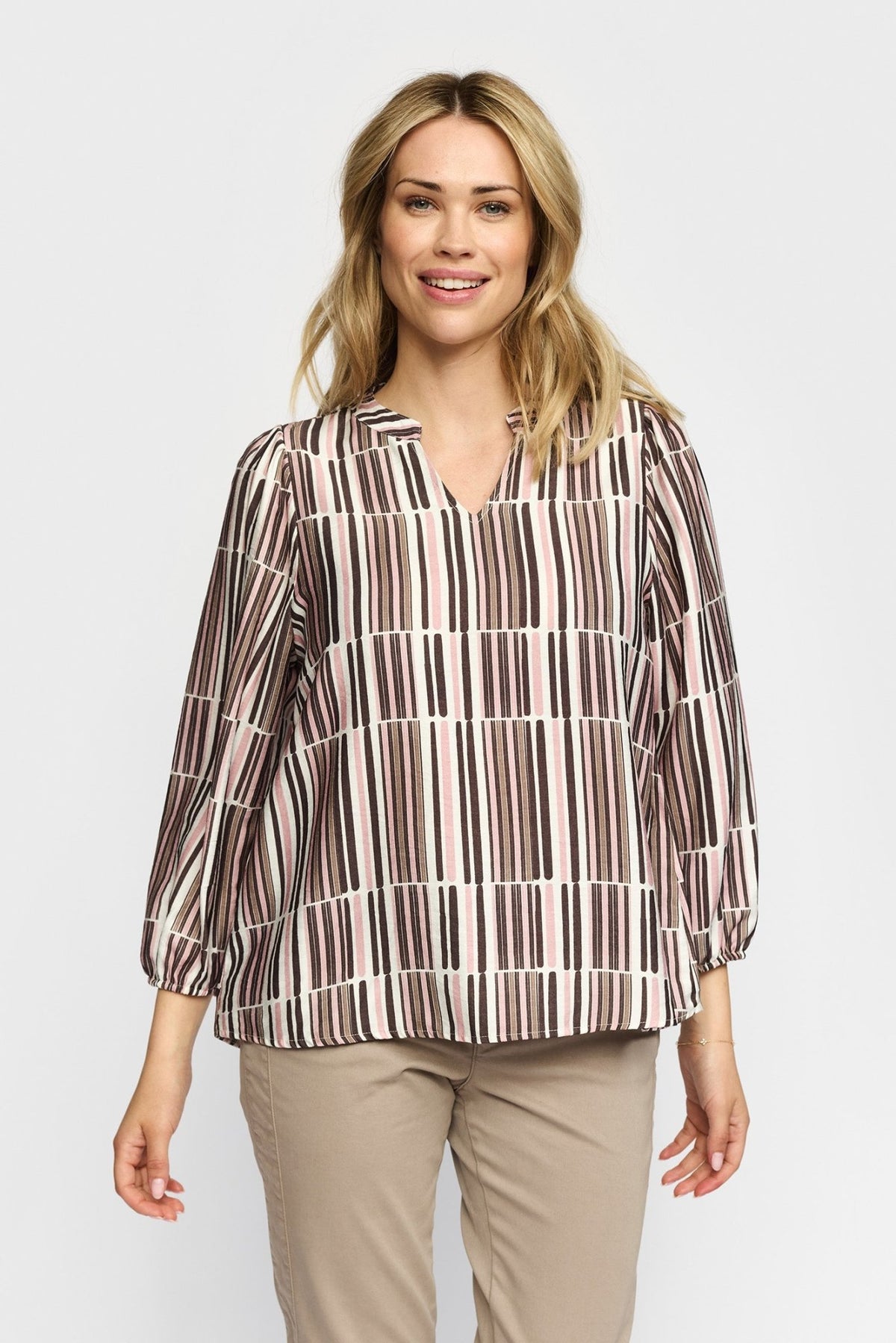 En smilende kvinde iført en 2-Biz Esmara Rosa-bluse med striber og beige bukser, der poserer foran en ensfarvet baggrund.