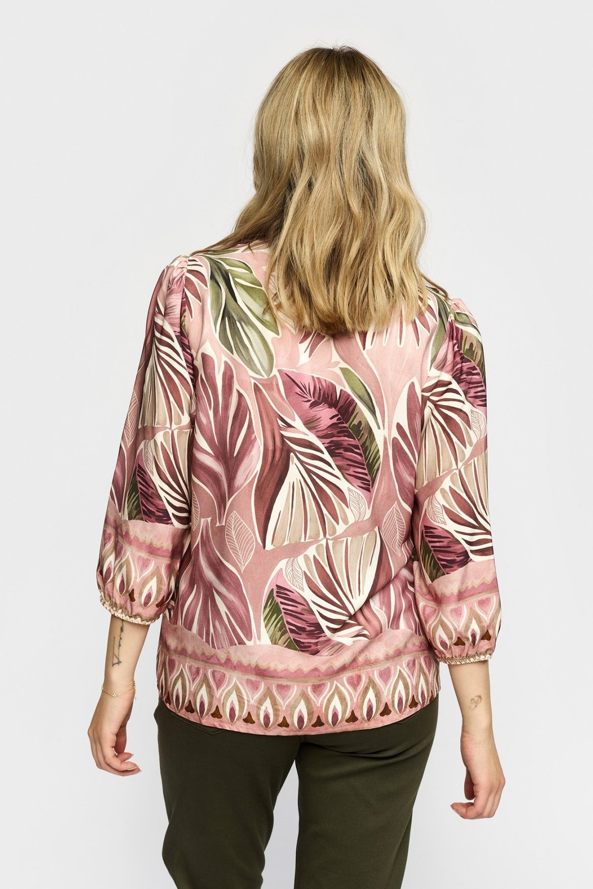 En blond person er iført en 2-Biz Ellen Gammelrosa-bluse og mørke bukser og ses her set bagfra foran en ensfarvet baggrund.