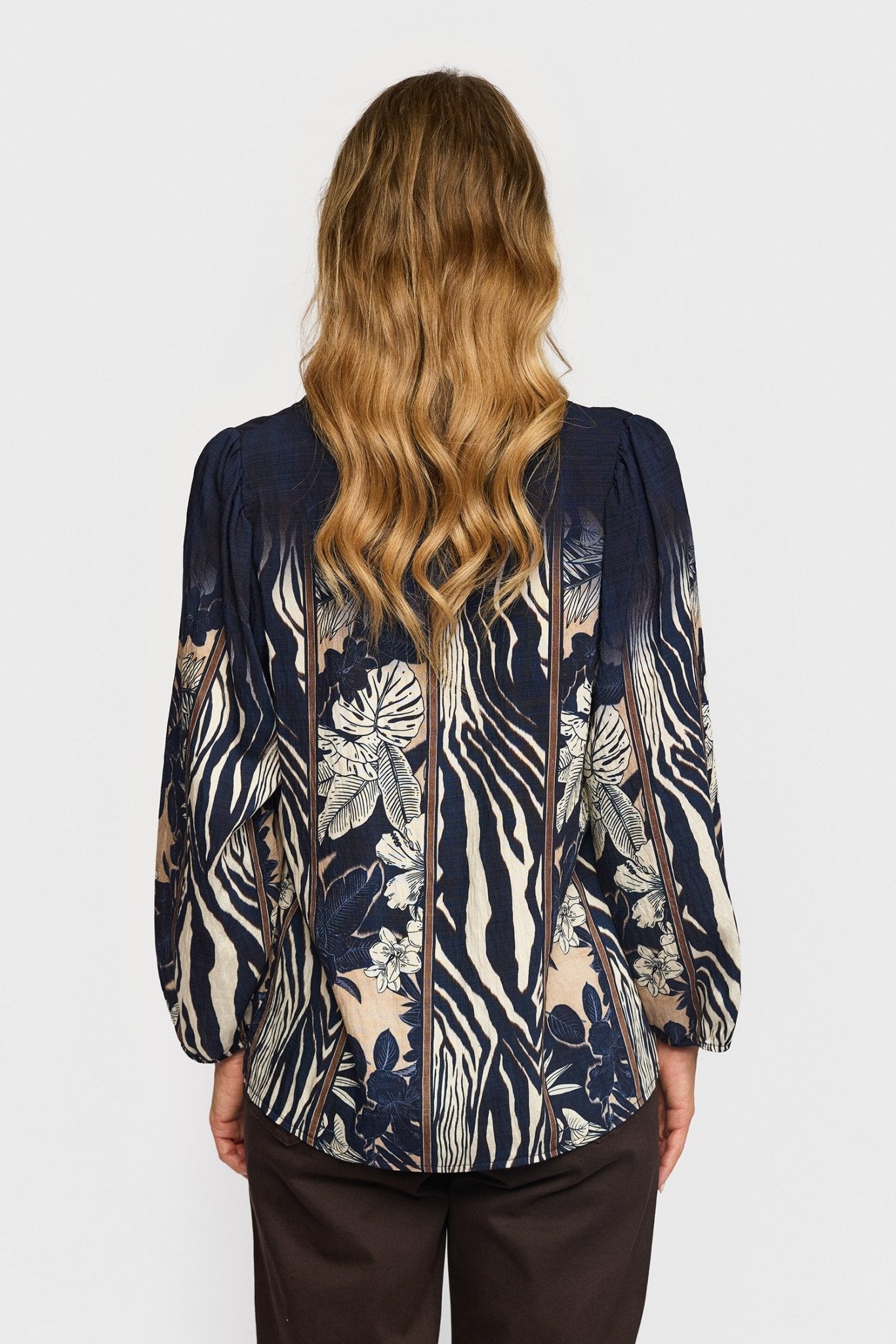 En person med bølget blondt hår iført en 2-Biz Lise Mørkeblå bluse med blomster- og zebraprint, set bagfra.