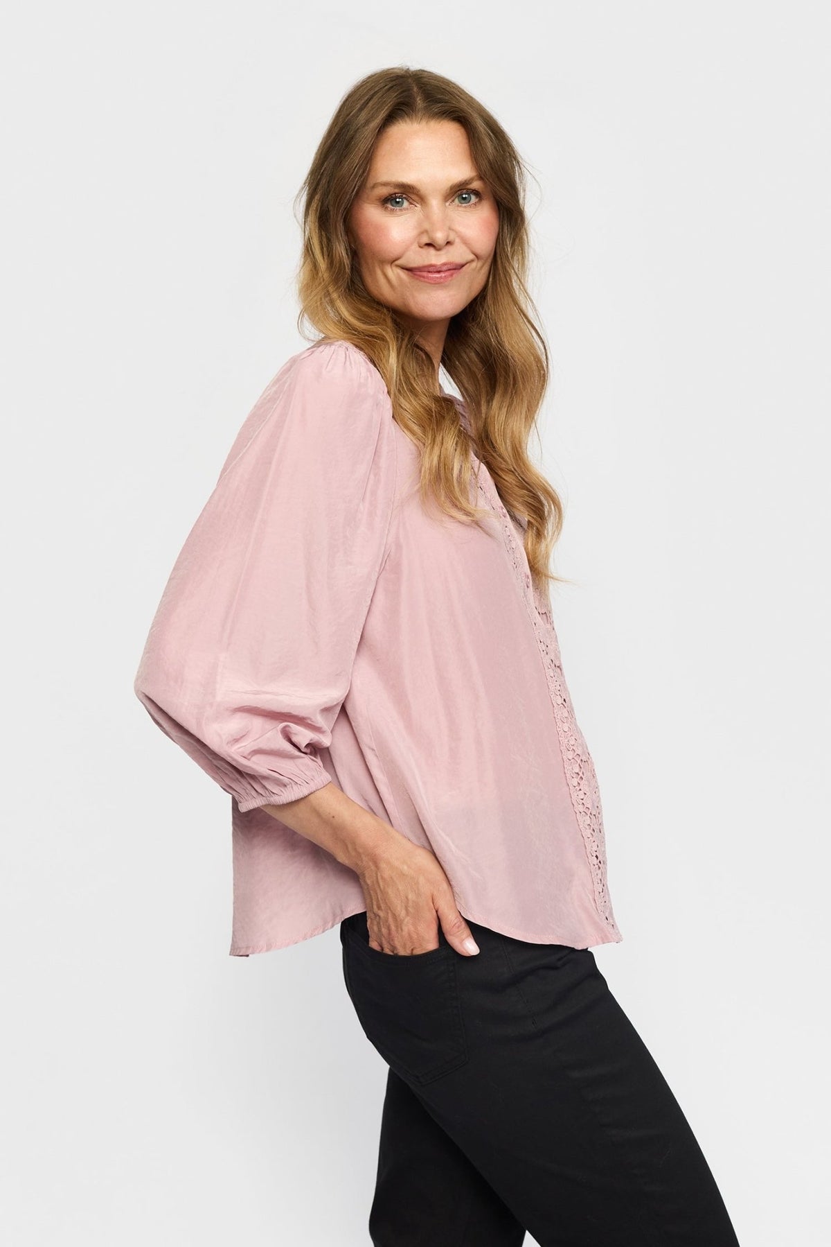 En kvinde med langt, bølget hår har en 2-Biz Karoline Rosa-bluse og sorte bukser på og står med siden til foran en ensfarvet baggrund.