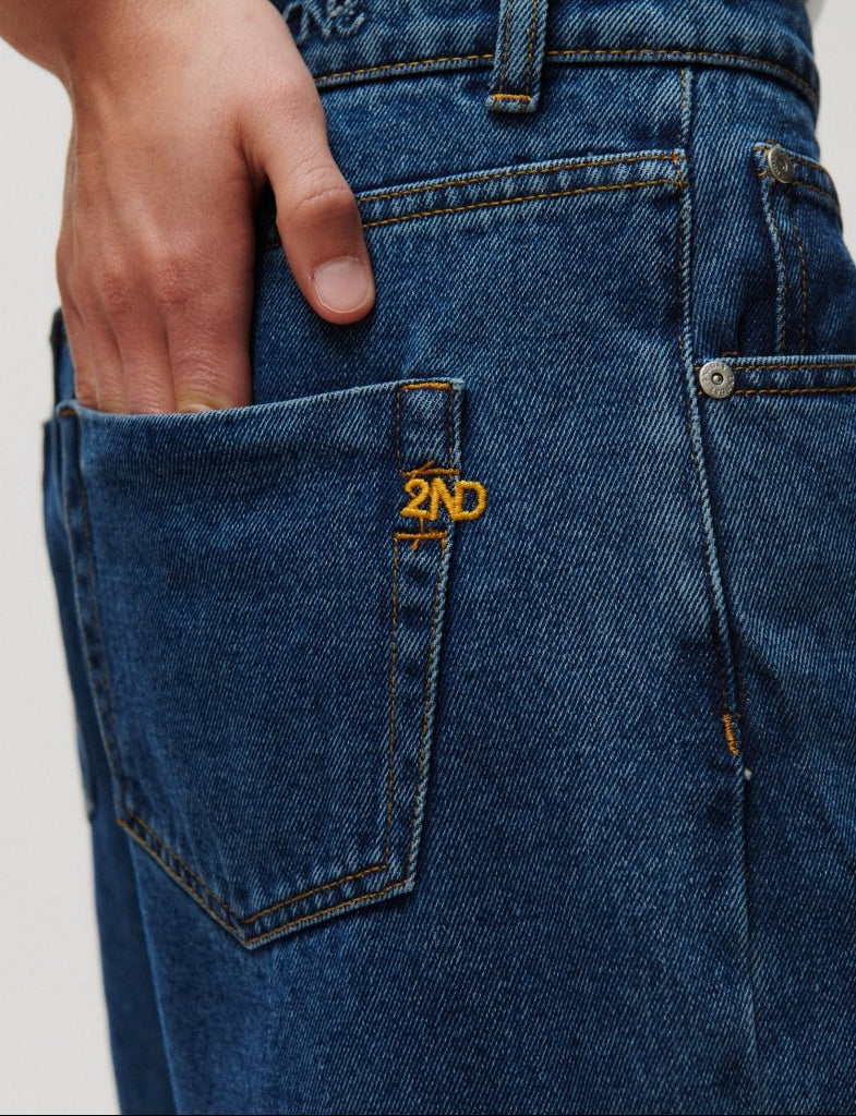 En hånd glider ned i baglommen på et par 2NDDAY Roden Denim-jeans i mellemblå, hvor der er broderet »2ND« over lommen.