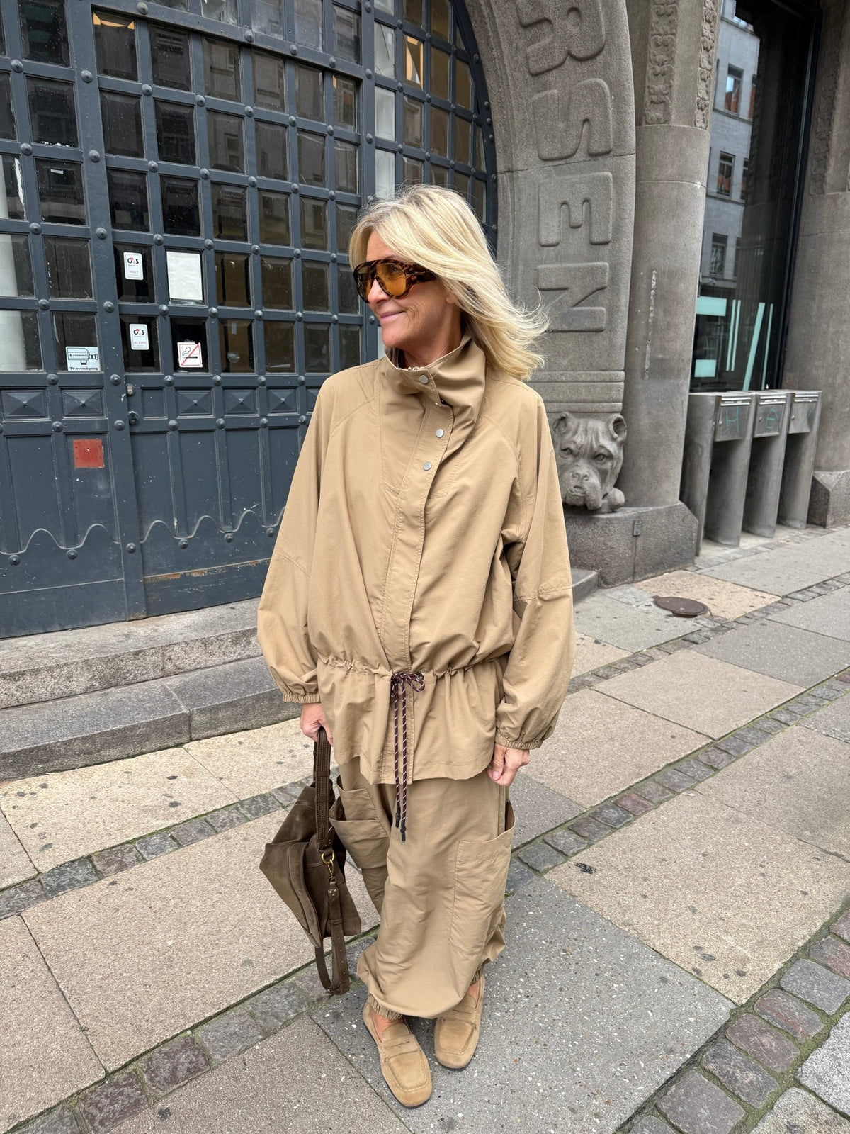 En kvinde iført en 2NDDAY 2NDKai-jakke og -bukser i farven »Camel« står på fortovet i byen med en brun taske og solbriller.