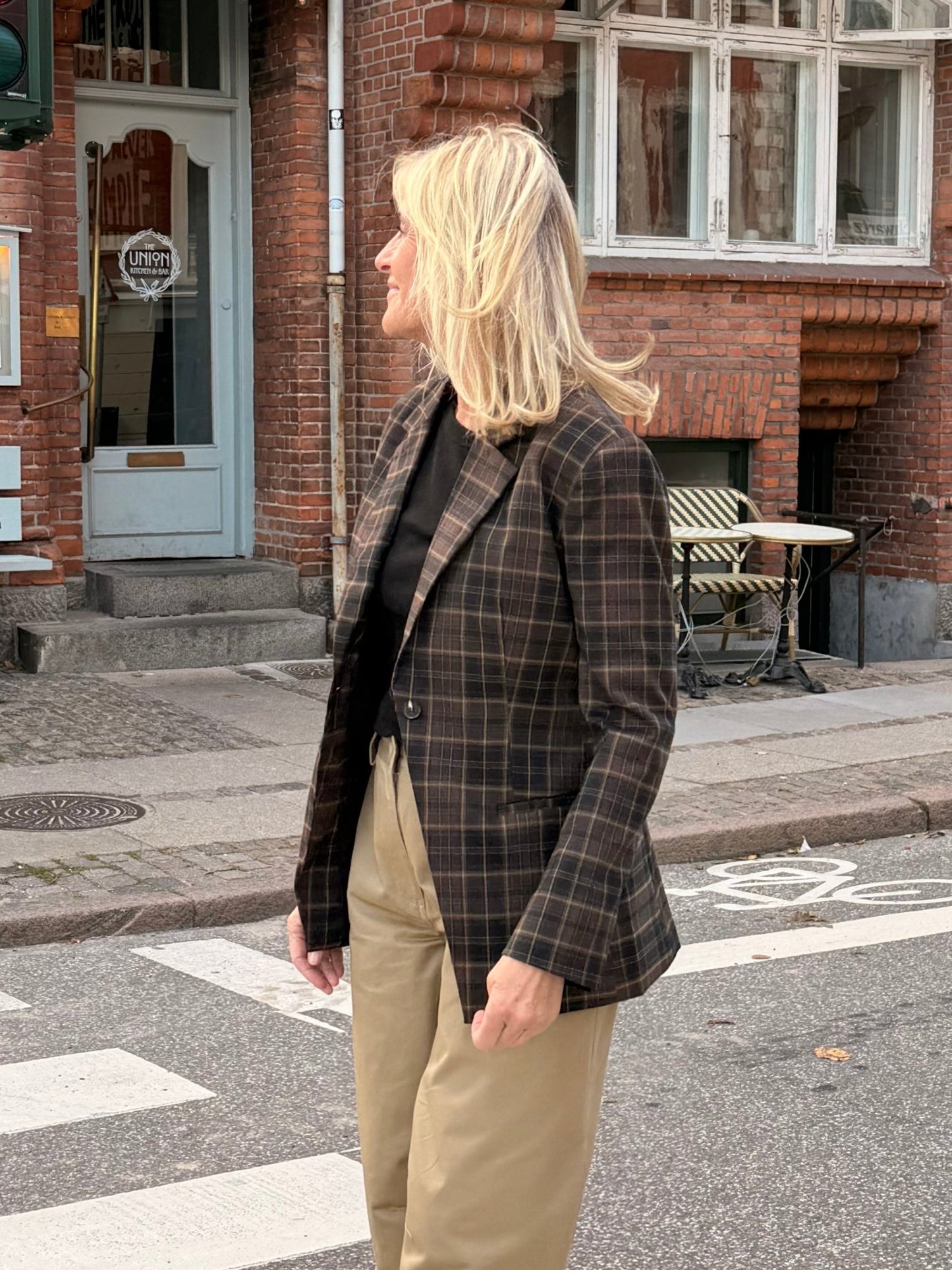 En kvinde med blondt hår, iført en ternet blazer fra 2NDDAY 2NDMaeva Ternet og kakifarvede bukser, står ved en café på en gade i byen.