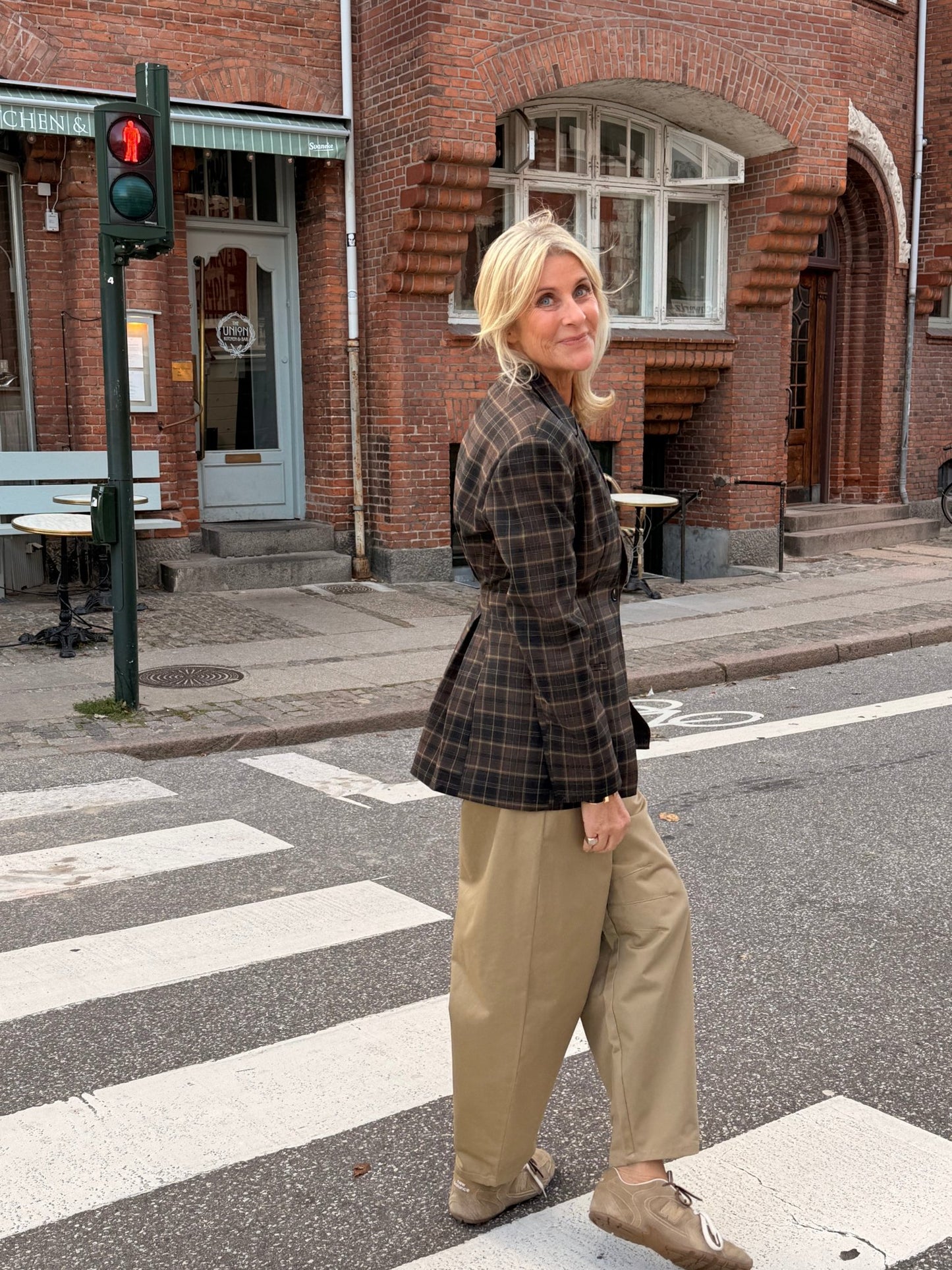 En kvinde iført en 2NDDAY 2NDMaeva Ternet-blazer i ternet mønster og khakifarvede bukser står ved nogle murstensbygninger under et grønt trafiklys.