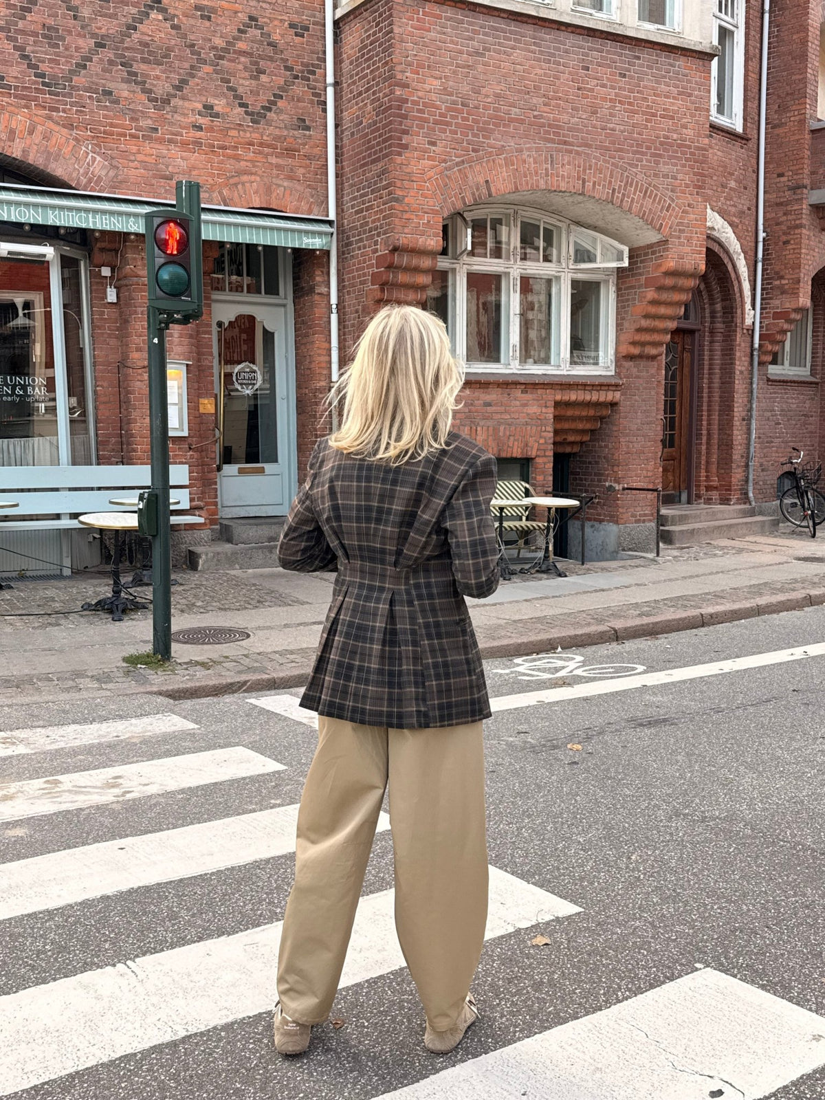 En person har 2NDDAY 2NDMaeva Ternet-blazeren i ternet mønster og beige bukser på ved et fodgængerovergang ved en murstensbygning.