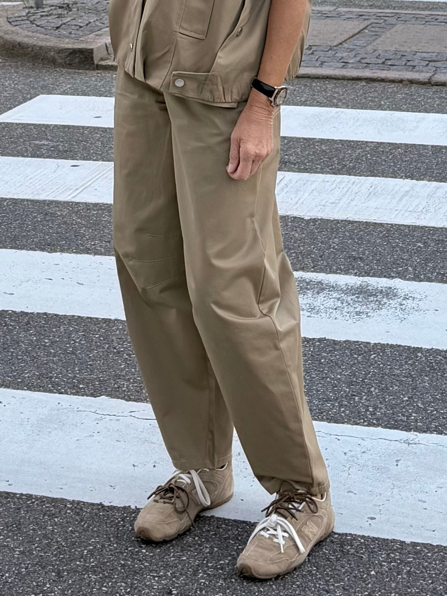 En person iført 2NDDAY 2NDJimmay Camel-bukser står på et fodgængerovergang med den ene hånd langs siden og har beige sneakers på.