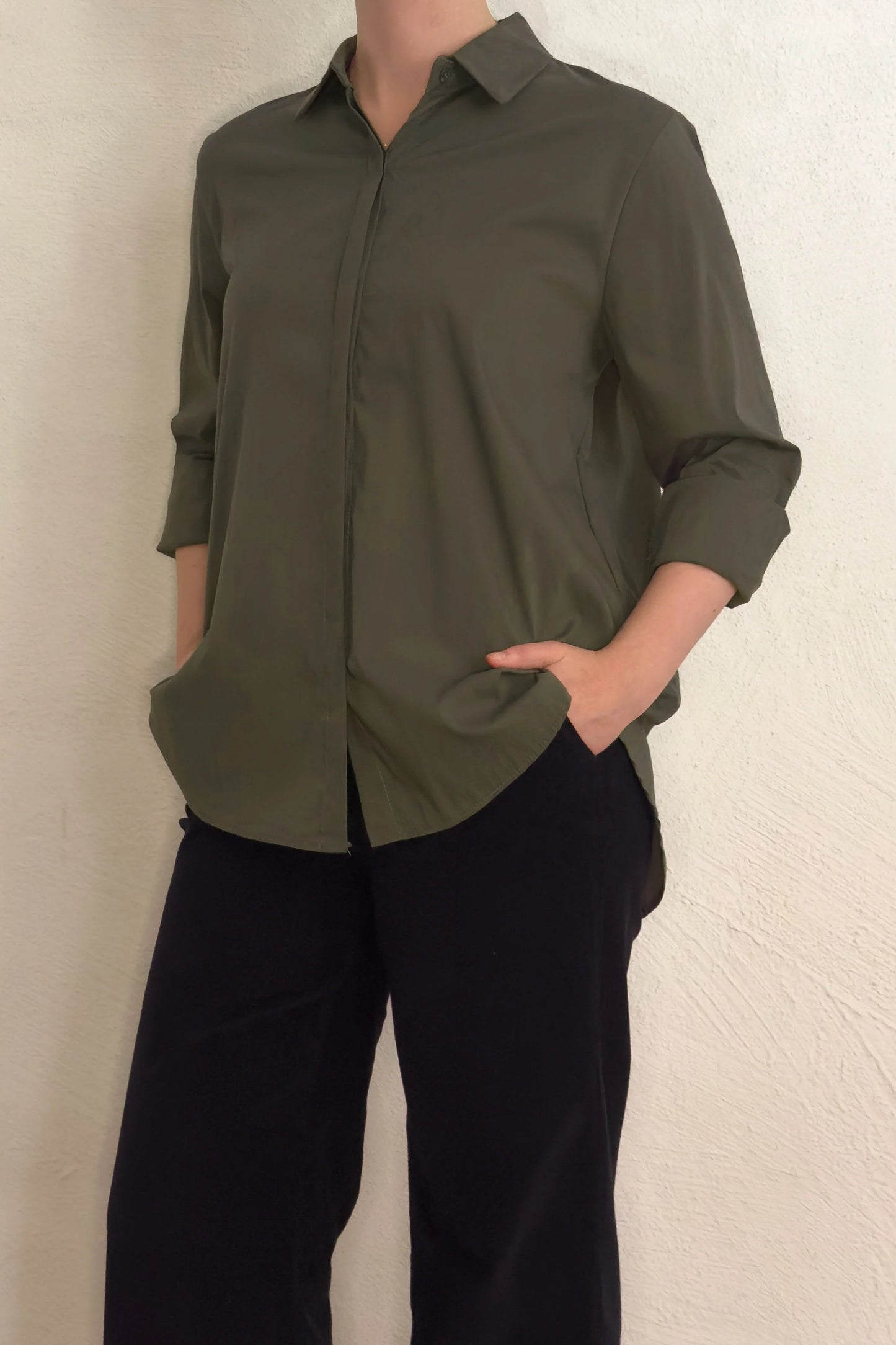 Model iført en grøn NORMA Army-skjorte fra Amaze Cph, med hænderne i lommerne og sorte bukser, foran en lys væg.