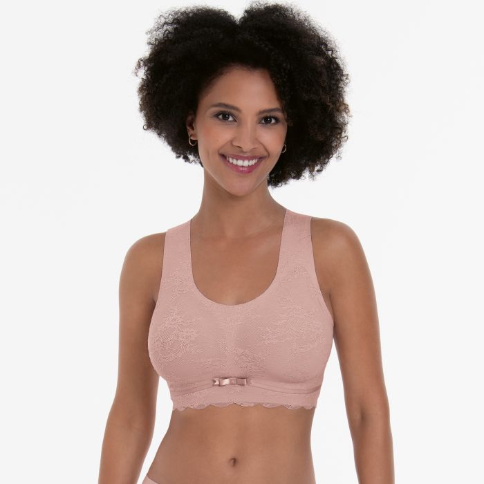 En kvinde iført en Anita Essential Lace Rosa-bralette, lyserød med sløjfe, står foran en ensfarvet baggrund.