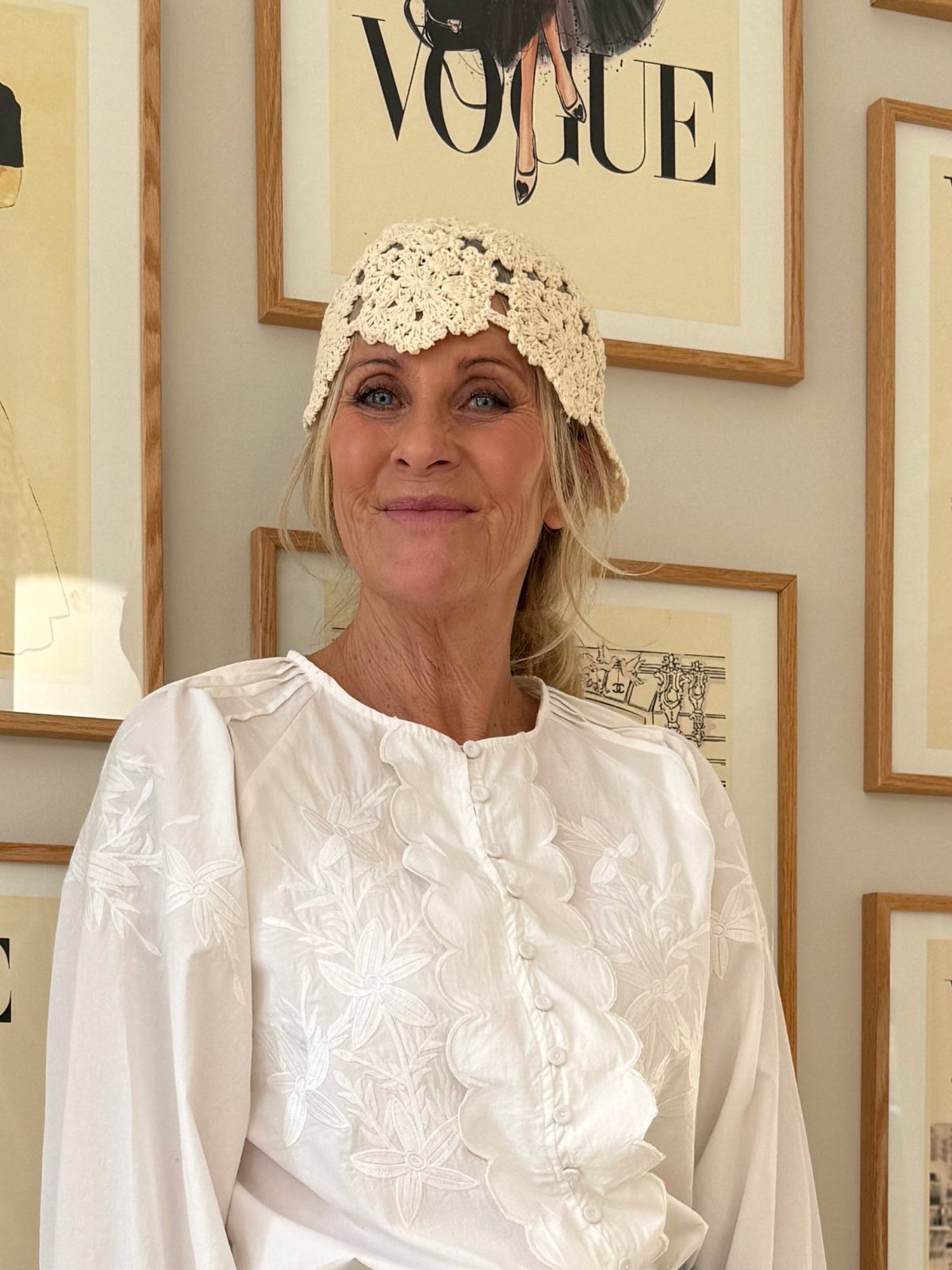 En kvinde iført en offwhite bluse fra Roseanne af mærket Black Colour og en vintagehat står ved siden af indrammede Vogue-forsider.