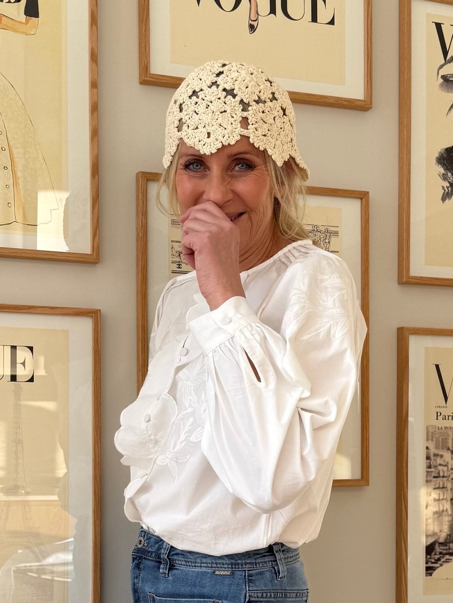 Roseanne fra »Woman in Black Colour« smiler i sin offwhite bluse foran indrammede Vogue-forsider, iført en vintage cremefarvet hat.