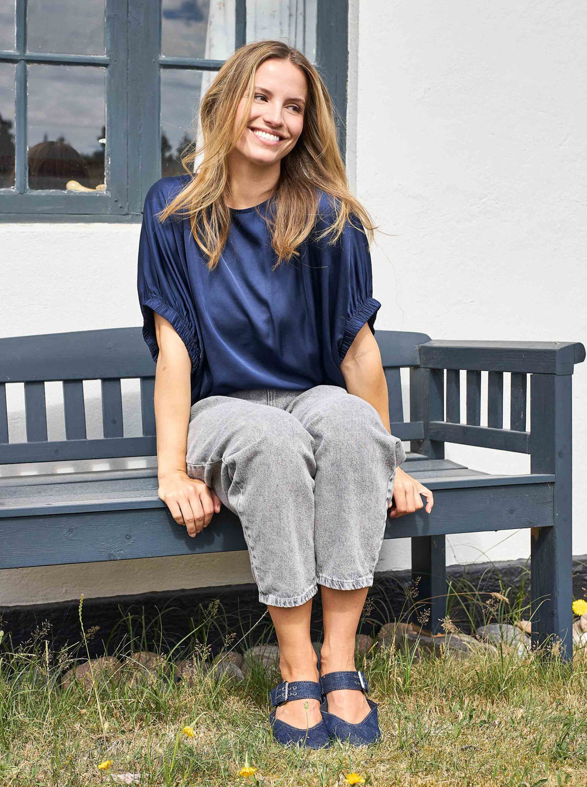 Kvinde med langt hår, der smiler udenfor, klædt i sort – Mabel Mørkeblå… bluse, grå bukser og blå sko.