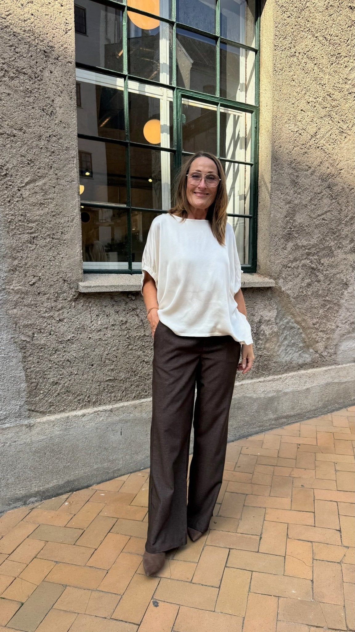 En kvinde er iført en sort Mabel-bluse i off-white og brune bukser på en murstensfortov ved en væg med struktur og et stort vindue.