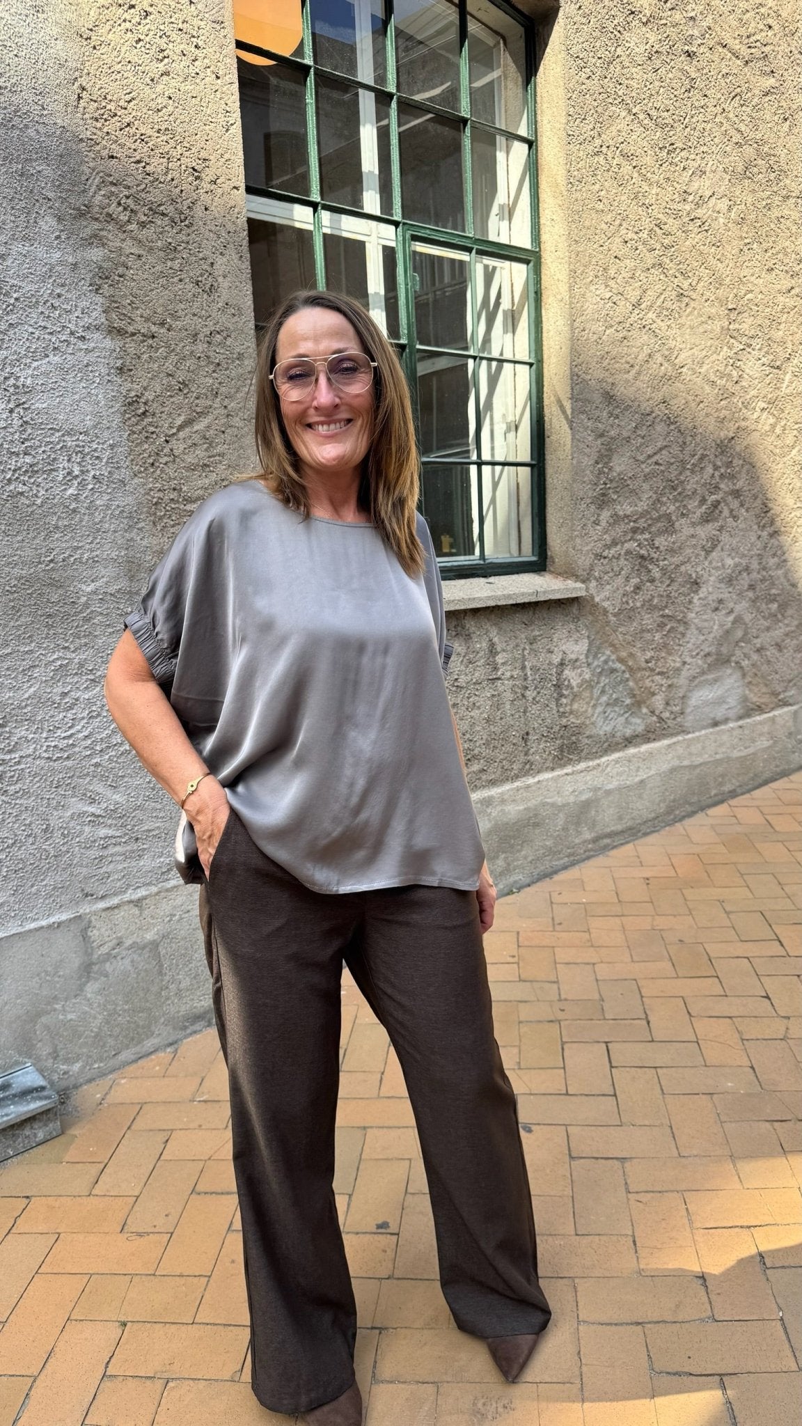 En kvinde udendørs på en murstensgang i en Mabel Mørkebrun-bluse fra Black Colour; hun smiler og ser afslappet ud.