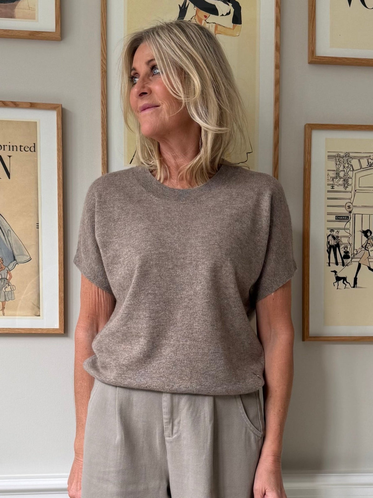 En kvinde iført en brunmeleret Vera Cashmere-trøje og beige bukser fra BTF CPH står ved siden af et indrammet kunstværk.