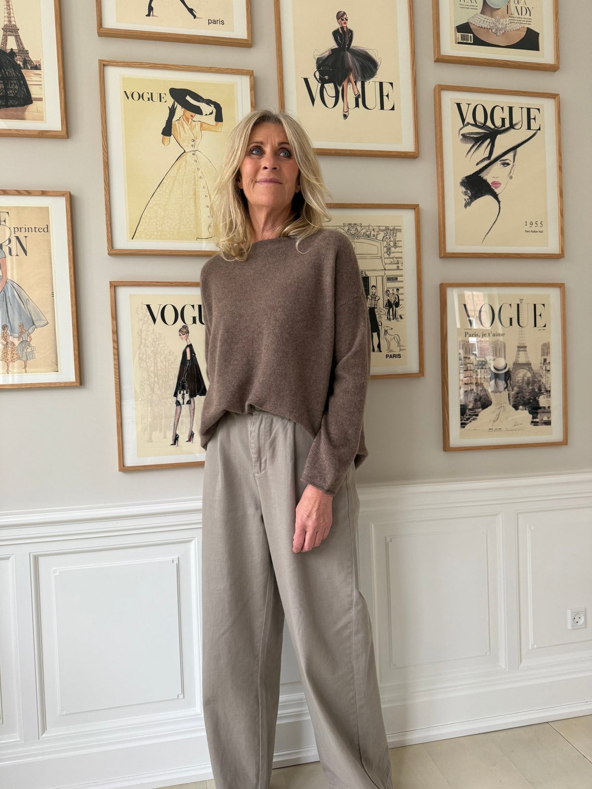 En kvinde iført en brun BTF CPH Mira Pure Cashmere-trøje står ved en væg med indrammede forsider fra magasinet Vogue.