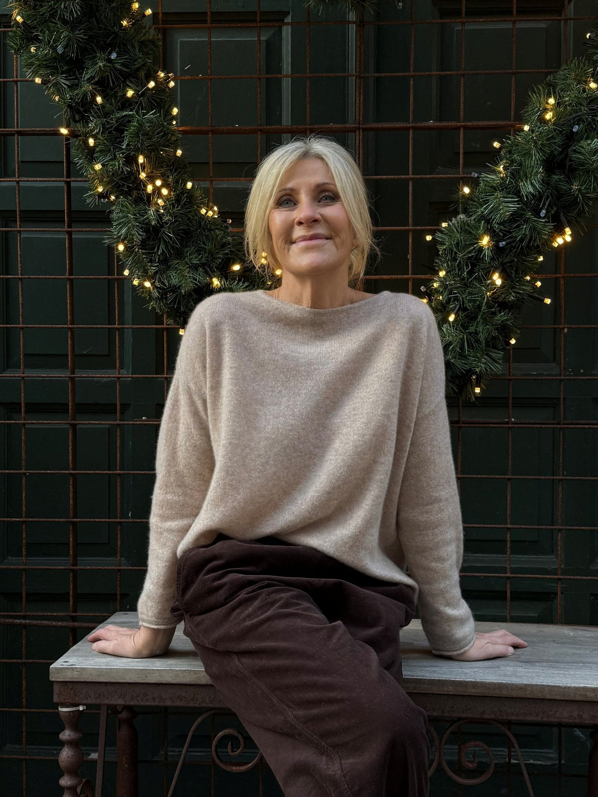 En kvinde i en lysebrun BTF CPH Mira Pure Cashmere 153490 sidder på en bænk foran en grøn væg med lyskæder.