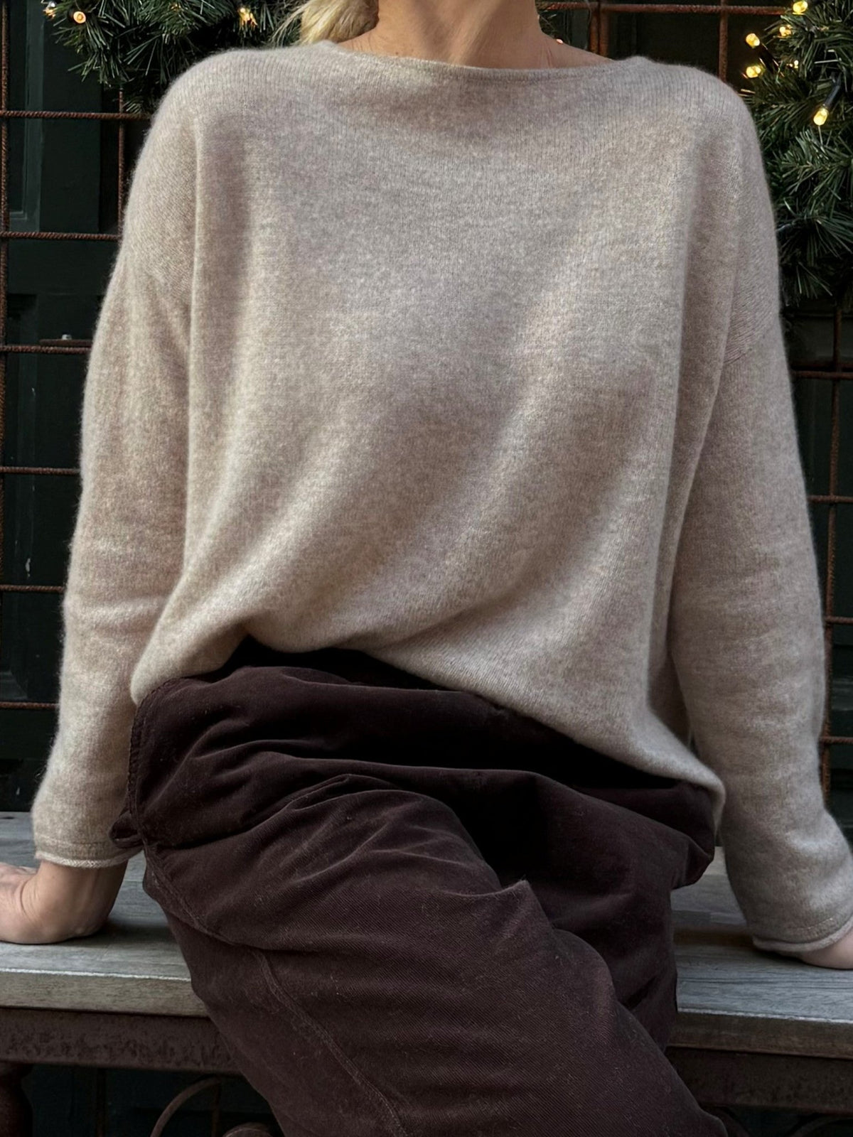 En person på en bænk iført en BTF CPH Mira Pure Cashmere Lysebrun-trøje og mørke bukser, med grønne planter og lys i baggrunden.