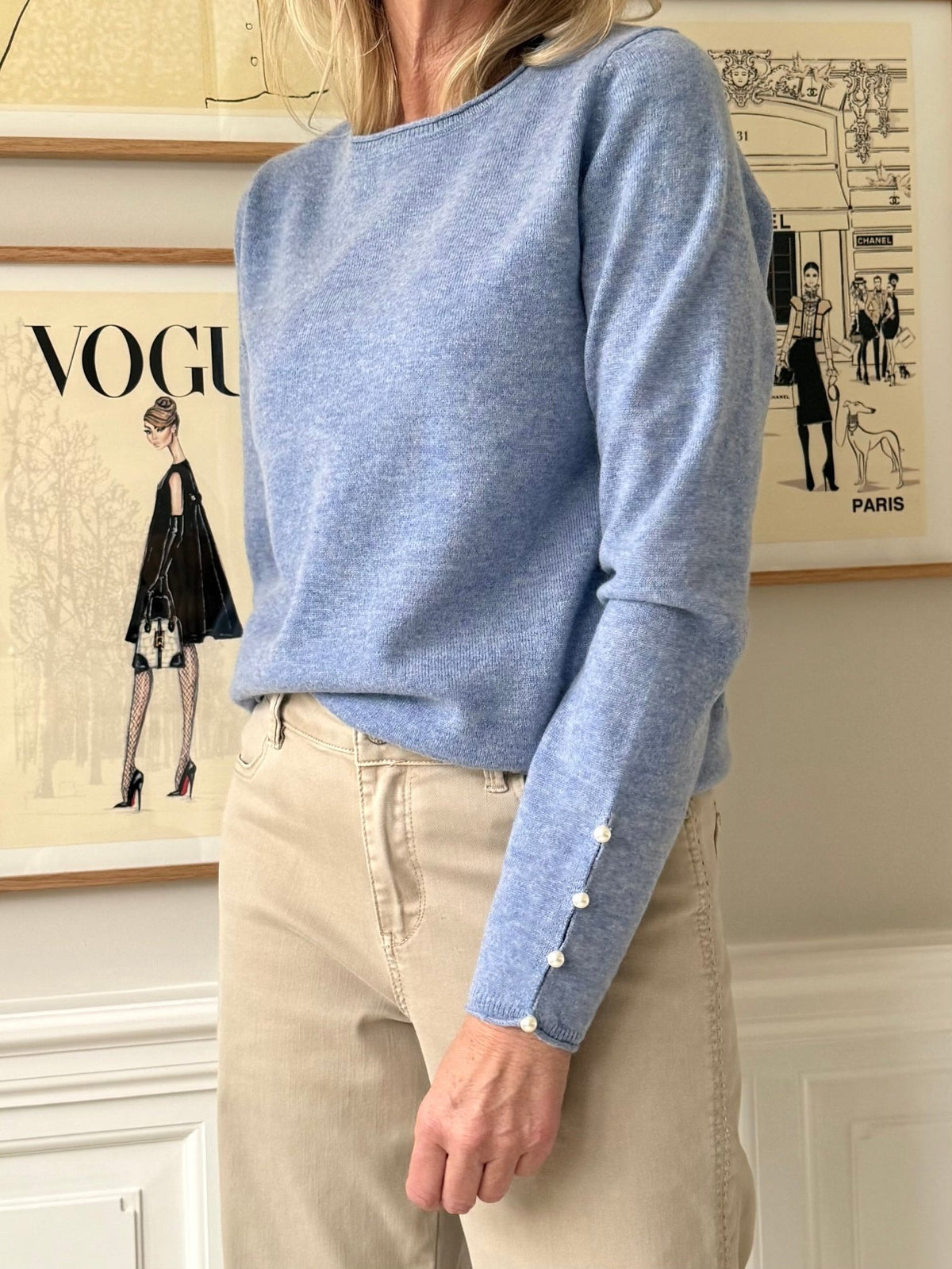 En kvinde iført en BTF CPH Cashmere Blå-trøje og beige bukser står foran indrammede modeillustrationer på væggen.
