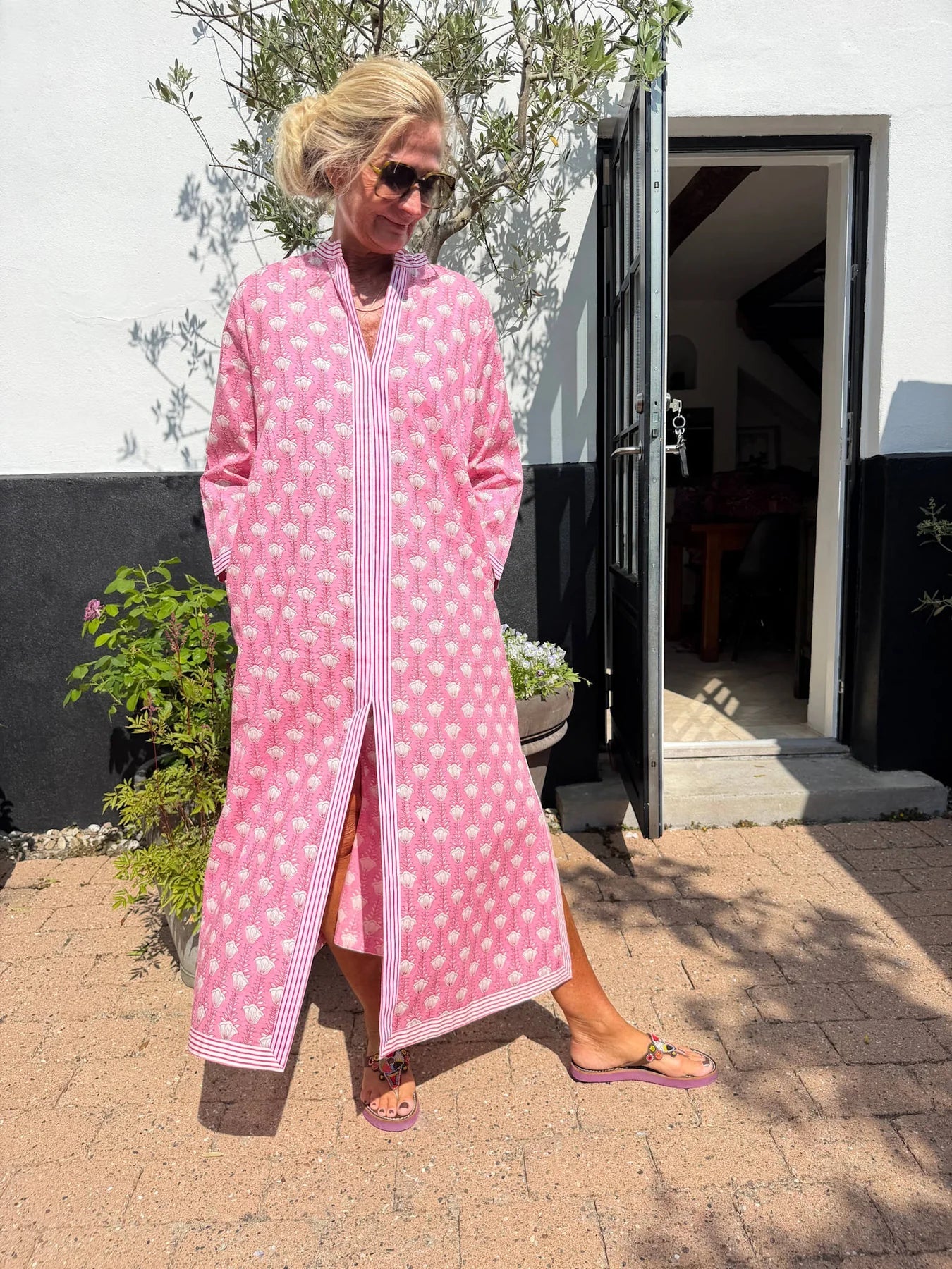 En kvinde i en lang, mønstret kjole fra Cabana Living – Zuri Pink – står udenfor ved en dør med hænderne i lommerne og blikket vendt nedad.