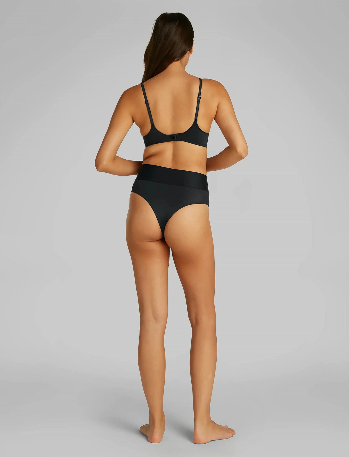En kvinde står med ryggen til, barfodet, iført Calvin Klein - Basic 2025 Black Sort, foran en ensfarvet baggrund.