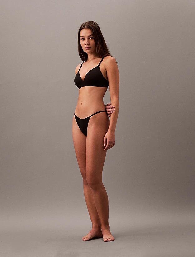 En kvinde iført en sort, trådløs Calvin Klein 150566-bikini, barfodet, med blikket rettet fremad foran en grå baggrund.