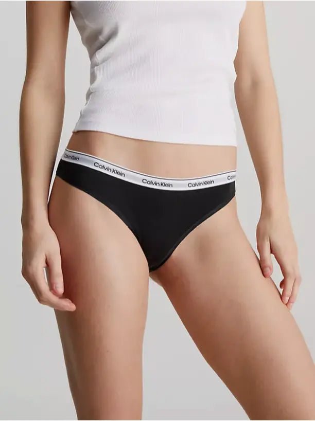 En person iført en hvid tanktop og sort Calvin Klein 153668-undertøj, der står foran en ensfarvet baggrund.