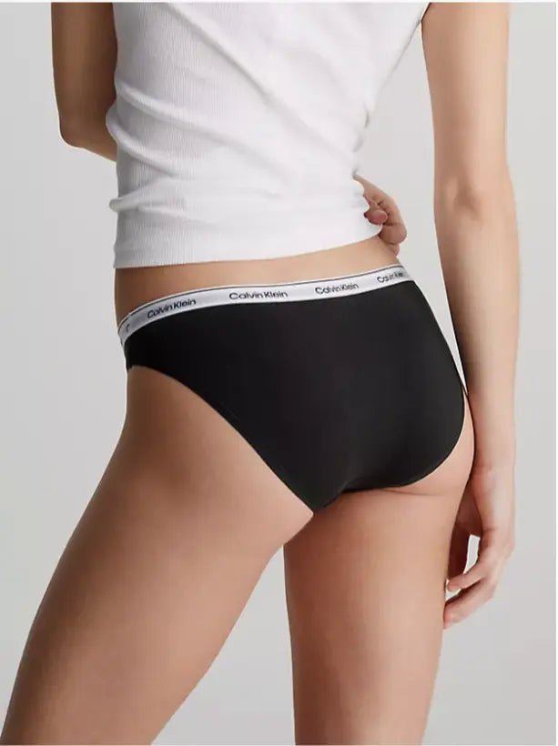 En person i en hvid tanktop og sorte Calvin Klein 3 FOR 399,- trusser, fotograferet bagfra foran en ensfarvet baggrund.