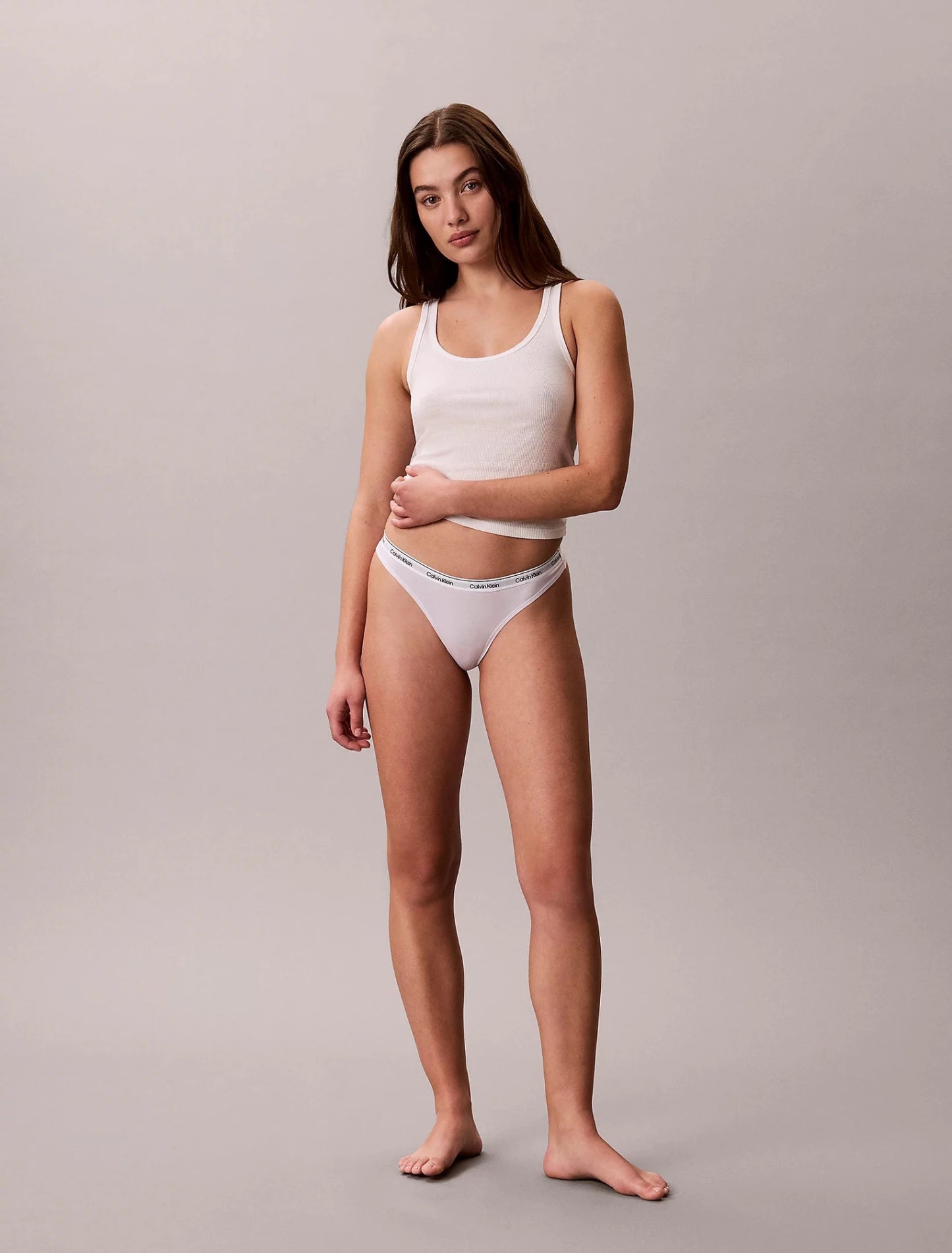 Kvinde i hvid tanktop og en Calvin Klein 3 FOR 399,- Lyselilla G-streng, barfodet, på en lysegrå baggrund.