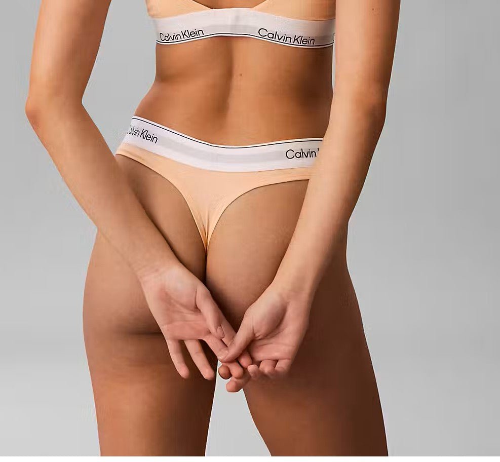 En person iført en Calvin Klein 4RW Fersken G-streng, med hænderne foldet bag ryggen, foran en ensfarvet baggrund.