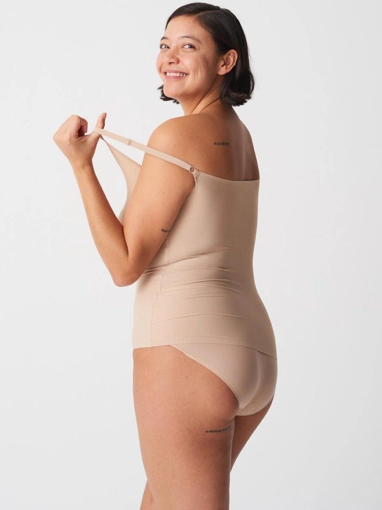 En smilende kvinde i en Chantelle Seamless Soft Stretch Camisole i farven Sand holder den ene skulderrem nede fra skulderen, på hvid baggrund.