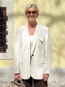 Kvinde i en off-white blazer fra Muse Tailor i København, med solbriller, udendørs ved en gul væg med et vindue til venstre.