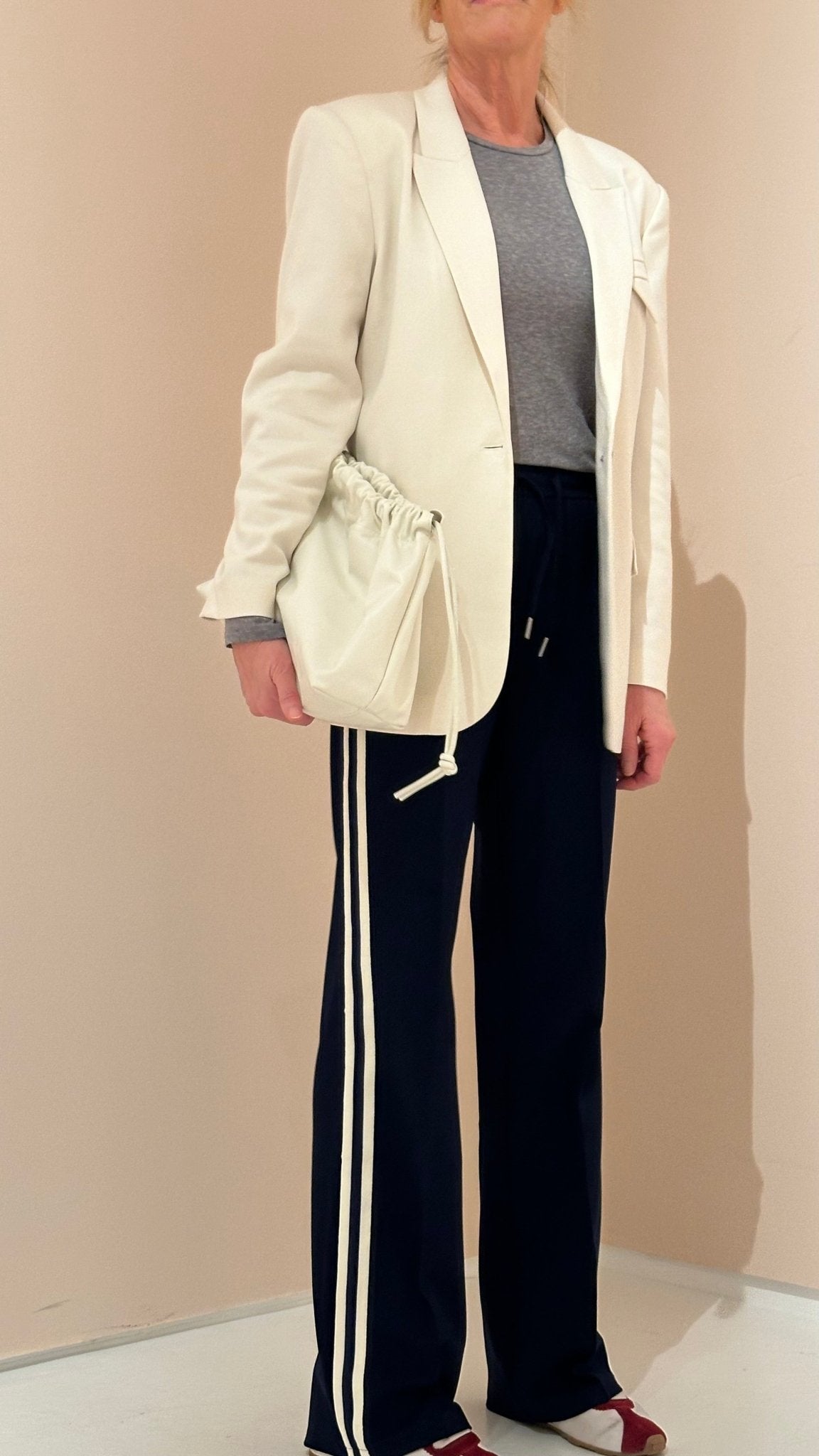 En person iført en off-white blazer fra Copenhagen Muse 140288, en grå skjorte og sorte bukser med hvide striber, der holder en hvid taske.