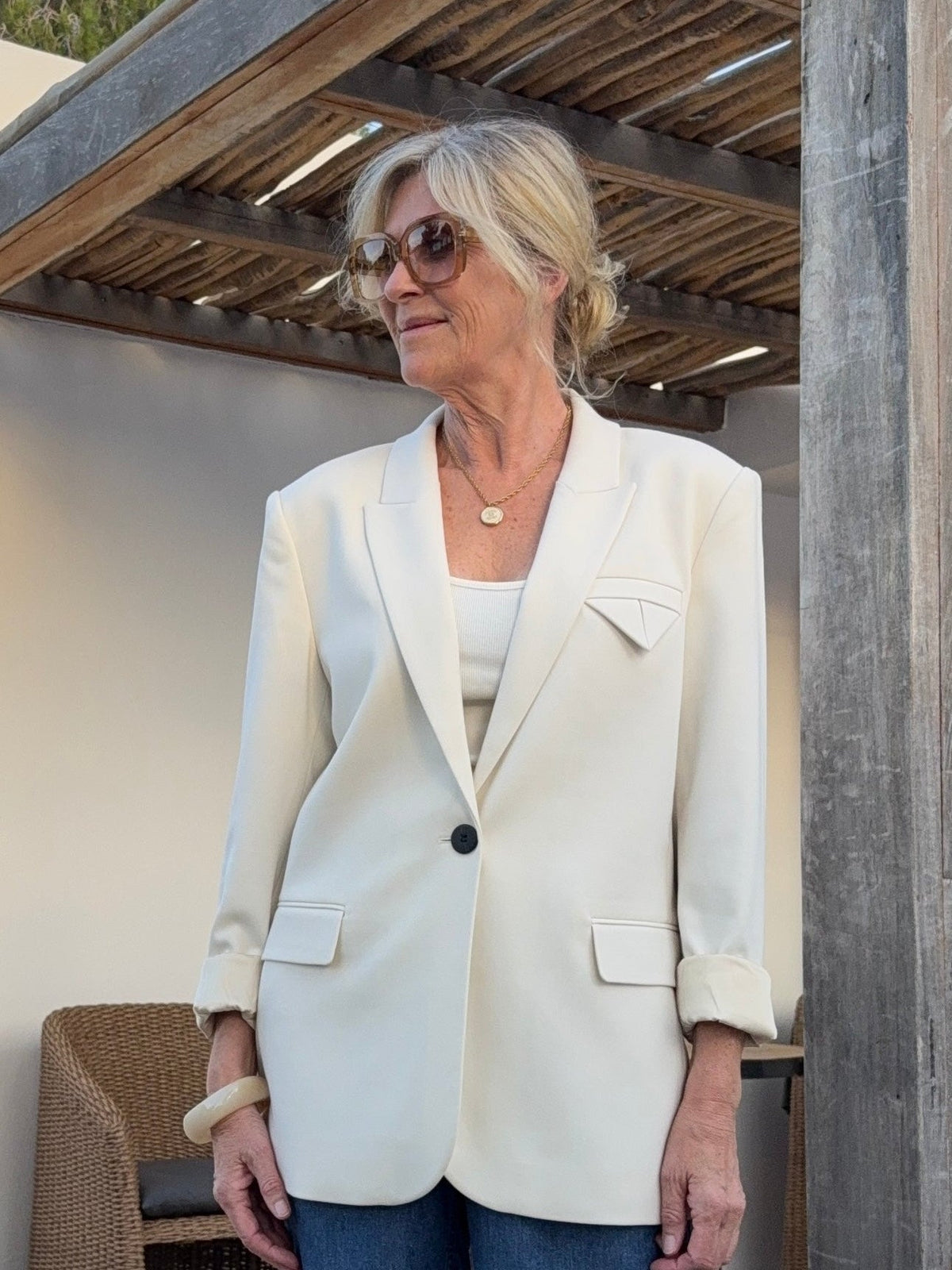En ældre kvinde i København, iført en off-white blazer fra Muse Tailor og solbriller, står udenfor tæt på kurvestole og træbjælker.
