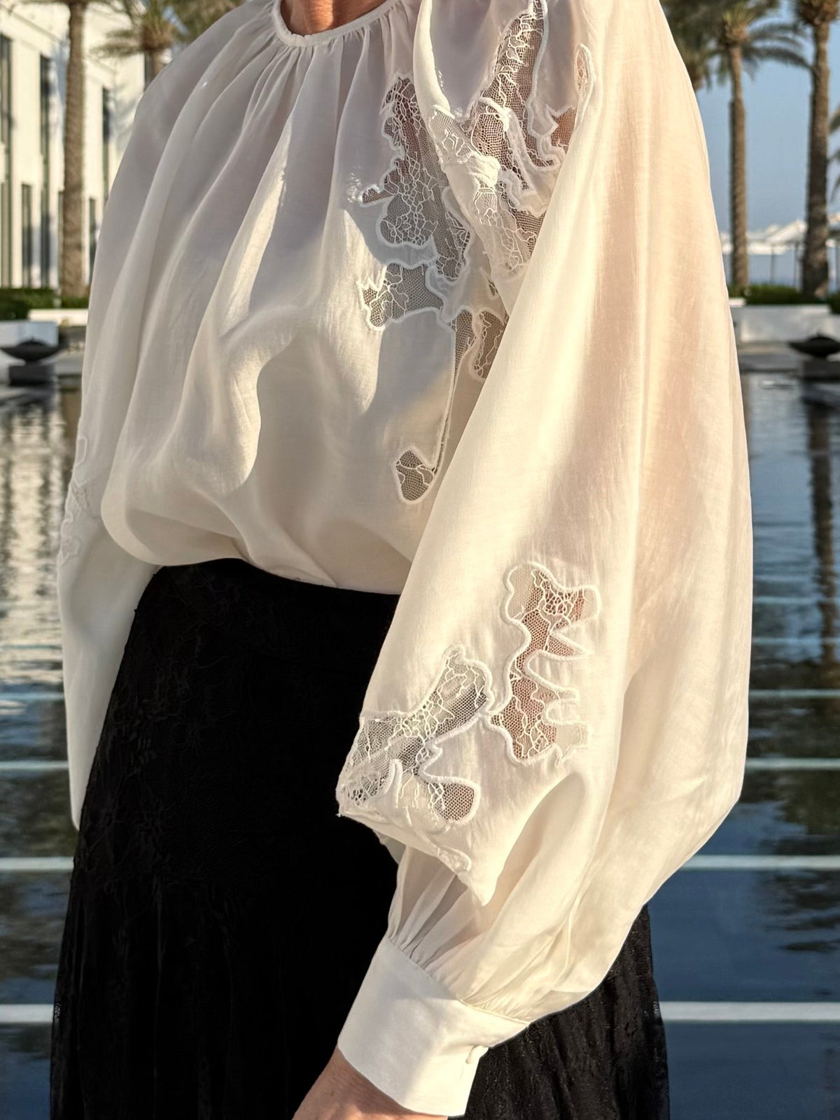 En person iført en Muse Molly-bluse i farven »Off White« med blonde-farvede ærmepatcher, tæt på vandet og med palmer i baggrunden.