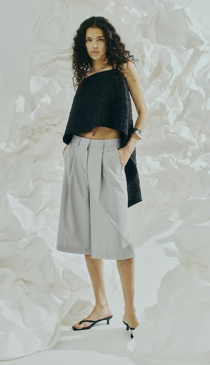 En kvinde iført en sort top, culottes fra Copenhagen Muse Tenna Lysegrå og sandaler står foran en krøllet hvid baggrund.