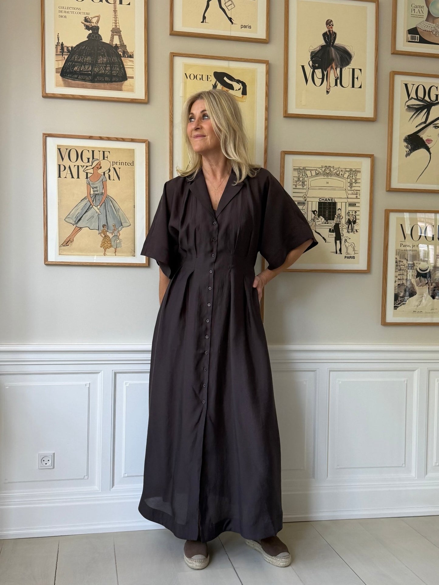 En kvinde iført en kjole fra Copenhagen Muse – Molly Mørkebrun står inde ved en væg med indrammede forsider fra magasinet Vogue.