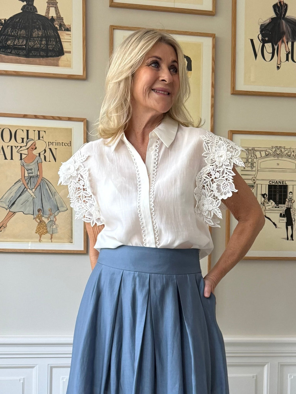 En kvinde iført en Muse Molly-skjorte i offwhite og en blå nederdel fra Copenhagen Muse står foran et indrammet modekunstværk på væggen.